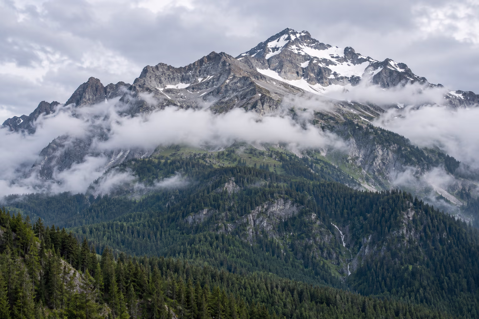 Cloud bands cross a mountain ridge between forested lower slopes and rocky high peaks.